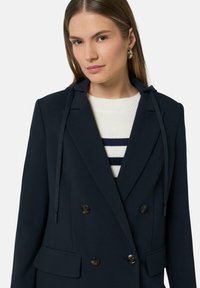 Marineblauwe dubbele blazer met capuchon, voorzien van twee grote knopen en een strakke uitstraling. Daaronder een wit sweater met marineblauwe strepen.