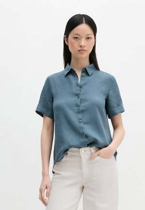 Kobieta ubrana w niebieską koszulę z krótkim rękawem wpuszczoną w jasnobeżowe spodnie z wysokim stanem, stojąca z jedną ręką w kieszeni.