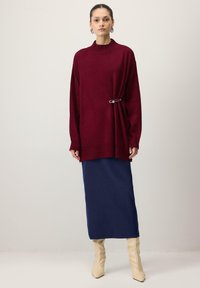 Pull oversize bordeaux avec un col montant, détail en boucle sur le côté, associé à une jupe longue bleu marine et des bottes crème montant jusqu'aux genoux.