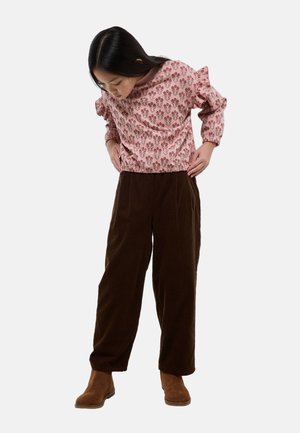 Fille portant un haut à manches longues à fleurs roses, un pantalon large marron foncé et des bottes marron, regardant vers le bas en ajustant sa tenue.