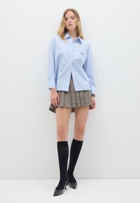 Camicia a righe blu chiaro con bottoni e maniche lunghe, abbinata a una gonna a pieghe a quadri marrone. Calze nere fino al ginocchio e tacchi borchiati completano il look.
