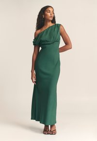 Zelena, svilnata, maxi obleka z enim ramenom z drapirano tkanino. Ima oprijet bodice in padajoče krilo, v kombinaciji z črnimi sandači z naramnicami.