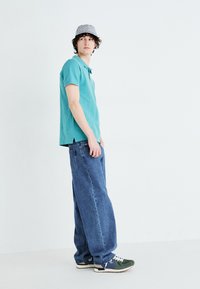 Camisa polo turquesa de mangas curtas, jeans azuis largos e ténis verdes. Boné com padrão preto e branco na cabeça. Fundo minimalista.
