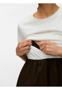 Personne soulève une chemise blanche pour révéler un soutien-gorge noir en dessous, portant une jupe brun foncé, sur un fond blanc uni.