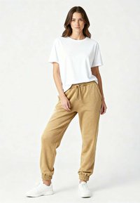 Femme debout portant un t-shirt blanc uni, un pantalon de jogging beige à cordon de serrage et des baskets blanches, sur un fond clair.
