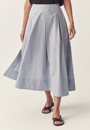 Jasnoniebieska spódnica midi w kształcie litery A z plisami z przodu, noszona z czarnymi, płaskimi sandałami na otwarte palce, na osobie o jasnej karnacji.