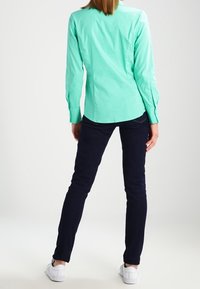 Camicia a maniche lunghe verde menta chiaro, design aderente, abbinata a jeans slim-fit blu scuro e sneakers bianche, vista da dietro.