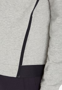 Grijze sweatshirt met zwarte zijpanelen en stiksels. Heeft een zachte textuur en een subtiel logo aan de onderkant aan de linkerkant.