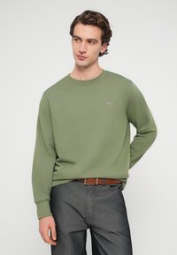 Jeune homme en sweat-shirt vert clair et ceinture marron portant un pantalon gris foncé, debout devant un fond blanc uni, regardant vers la gauche.