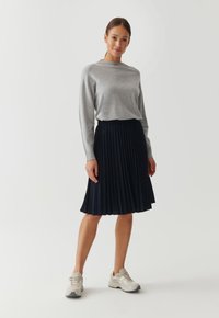 Chemise grise à manches longues associée à une jupe plissée navy. L'ensemble présente une coupe décontractée et une longueur au genou, complété par des baskets beige.