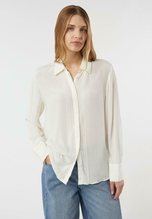 Lightweight, cream blouse with a collar, button placket, and vertical pleats. Features long sleeves and a relaxed fit, paired with blue jeans.