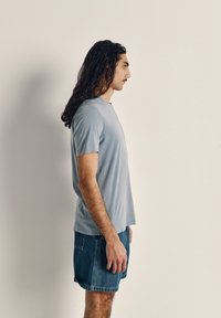Light blue cotton t-shirt with short sleeves, round neckline, and relaxed fit, paired with blue denim shorts. Side view of the model.