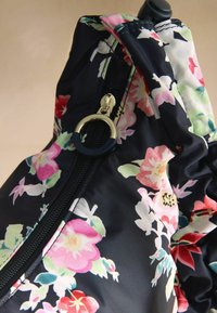 Musta kangas värvilise lillemustriga, mille keskmes on ümmarguse kuldse ja musta tõmbelukuga tõmblukk.