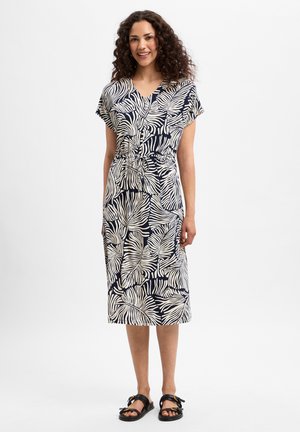 Frau steht lächelnd da, trägt ein knielanges schwarz-weißes Kleid mit Blattmuster, Kurzarm und schwarze Sandalen.