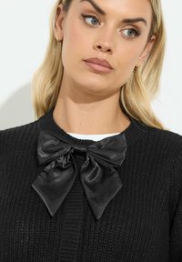 Svarttexturerad kofta med en satin svart rosett i halsen. Designen har ett ribbat mönster och en enfärgad yta med rund hals.