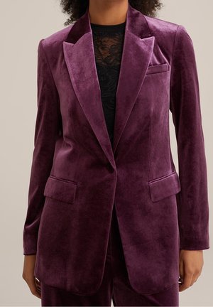 Blazer en velours violet avec une texture douce, design à boutonnage simple, revers crantés et poches latérales. Présente une coupe ajustée.