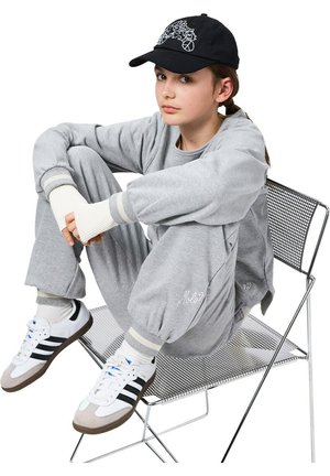 Young person sitting on metal chair with knees up, wearing grey tracksuit, white Adidas sneakers, and black cap with white design.