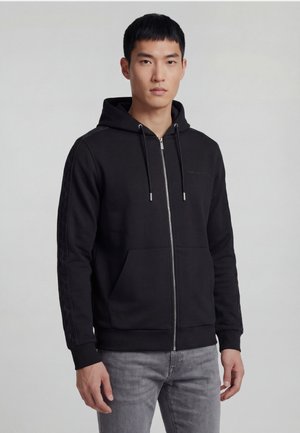 Young man wearing a black zip-up hoodie with drawstrings and side stripe detail, paired with light gray jeans, standing against a plain background.