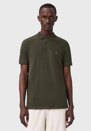 Homme portant un polo à manches courtes vert olive avec trois boutons et un pantalon blanc, debout devant un fond gris clair uni.