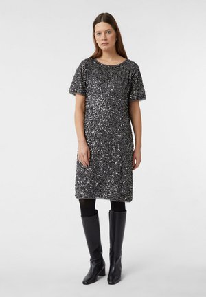 Una donna indossa un vestito argento con paillettes, di lunghezza ginocchio e maniche corte, abbinato a collant neri e stivali al ginocchio in pelle nera, mentre si trova di fronte a uno sfondo neutro.