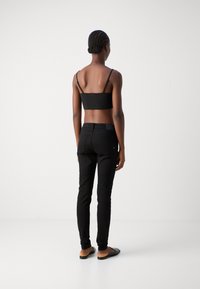 Mujer con trenzas llevando un top negro corto, jeans ajustados negros y mocasines negros de lona, de pie frente a un fondo liso.