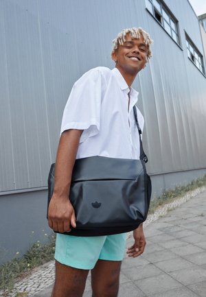 Młody mężczyzna z jasnymi blond dredami, ubrany w białą koszulę i miętowe szorty, noszący czarną torbę na ramię na zewnątrz obok szarego budynku.