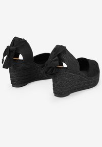 Black wedge sandals featuring a textured woven heel, soft suede upper, and decorative bows at the back for added detail.