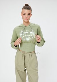 Sudadera recortada de color verde oliva con texto gráfico blanco, mangas largas, cordón ajustable, combinada con pantalones baggy beige.