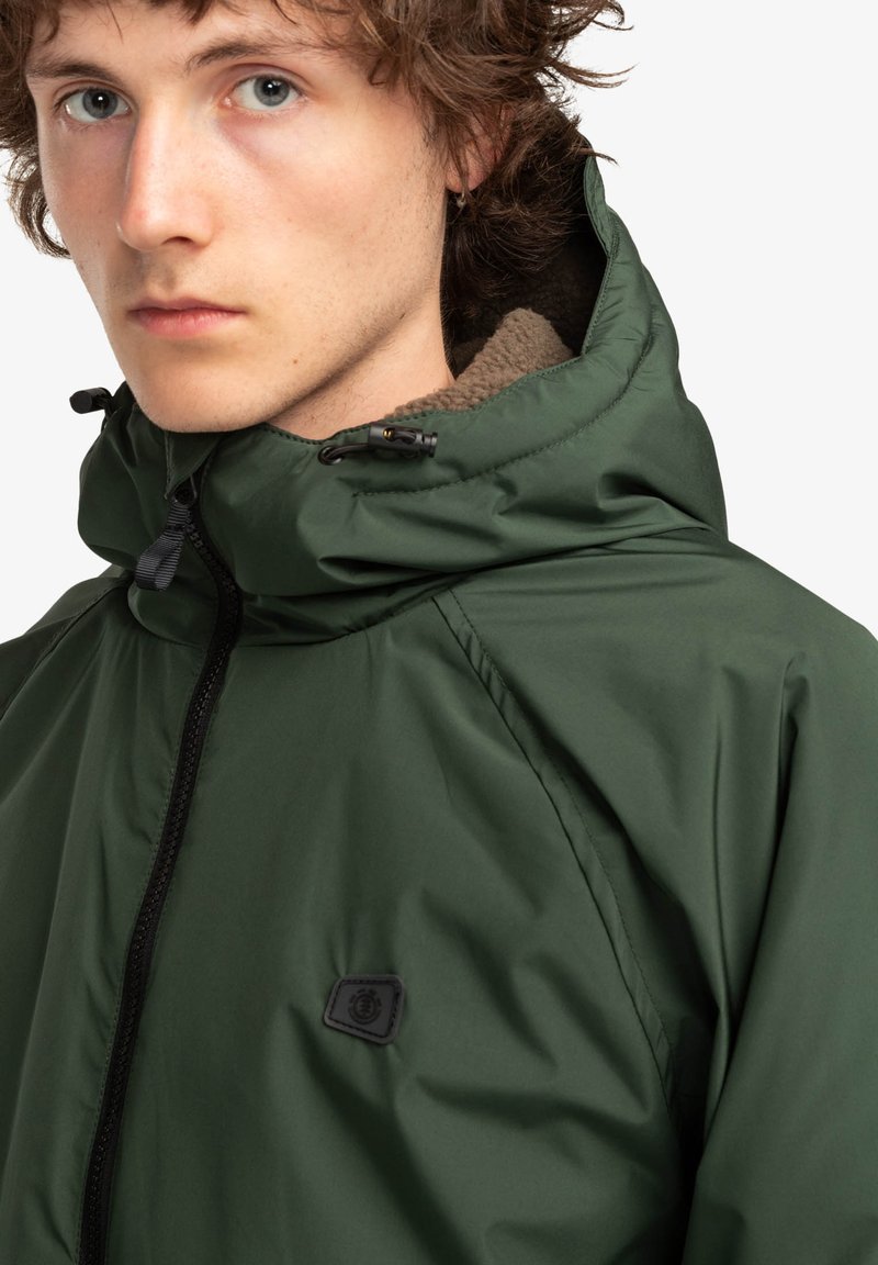 Giacca verde con cappuccio, dotata di zip frontale, fodera interna morbida e piccolo patch con logo nero sul petto. Tessuto liscio e resistente all'acqua.