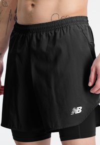 Primer plano de una persona que lleva pantalones cortos deportivos negros con mallas de compresión incorporadas, mostrando parte del torso tatuado y las manos.