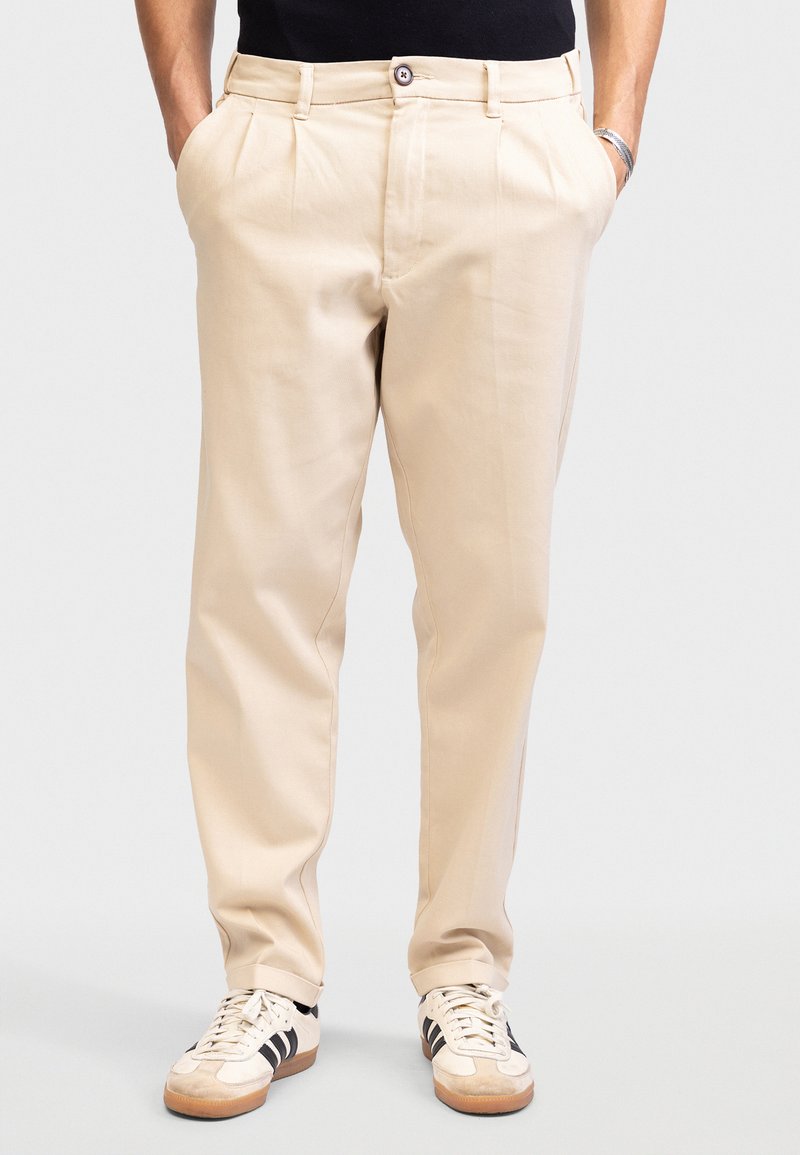 Man draagt beige geplooide broek met handen in de zakken en witte sneakers met zwarte strepen, staand tegen een effen achtergrond.