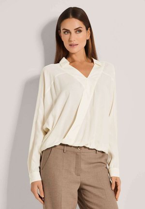 Crèmekleurige blouse met een V-hals en lange mouwen, met een losse pasvorm en een ingestopte zoom. Gecombineerd met lichtbruine maatpakken.