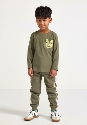 Jonge jongen staat, gekleed in een olijfgroen shirt met lange mouwen en een cartoondierenpatch, en bijpassende trainingsbroek met witte strepen en sneakers.