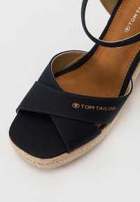 Tummansininen kangas-hiekkaliukas sandaali ristiinnaulituilla hihnoilla, luonnonsisustuksettua köyttä pohjan ympärillä ja kultainen "TOM TAILOR" -logo yläosassa.