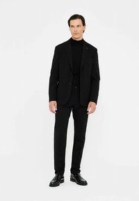 Abito nero con blazer aderente e pantaloni coordinati, abbinati a un dolcevita nero. Scarpe nere eleganti completano il look.