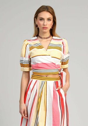 Jeune femme portant un haut cache-cœur rayé coloré à manches bouffantes et une jupe plissée assortie, main dans la poche, fond neutre.