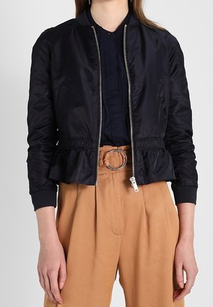 Vrouw draagt een zwarte bomberjack met ritssluiting over een donkere blouse, gecombineerd met een hooggetailleerde beige broek met een ronde metalen gesp.