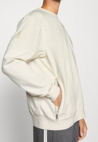 Lys beige sweatshirt med rund halsudskæring, lange ærmer, sidelomme med lynlås og struktureret stof. Har et diskret logo på ærmet.