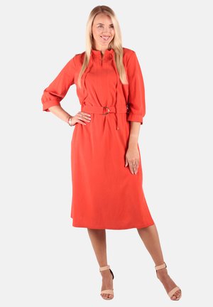 Blonde Frau lächelt, trägt ein knielanges orangefarbenes Kleid mit Dreiviertelärmeln und Gürtel, kombiniert mit nudefarbenen offenen High-Heels.