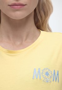 Κοντινό πλάνο ενός ατόμου που φοράει κίτρινο πουκάμισο με τη λέξη "MOM" κεντημένη, με ένα λουλούδι να αντικαθιστά το γράμμα "O" στο στήθος.