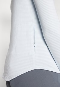 Helles blaues Longsleeve-Oberteil mit geripptem Textur, seitlichen Schlitzen und sichtbarem Taschendetail. Der Saum ist gebogen und betont einen geschichteten Look.