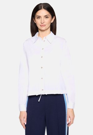 Frau mit glattem dunklem Haar, die ein weißes Hemd mit Knöpfen und marineblaue Hosen mit hellblauen und beigen Seitenstreifen vor einem schlichten Hintergrund trägt.