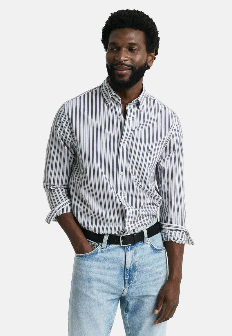 Chemise à rayures dans les tons blanc, marine et bleu clair ; avec un col classique et des manches retroussées, assortie à un jean bleu clair et une ceinture noire.