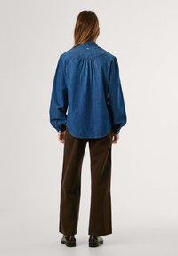 Denimhemd mit langen Puffärmeln, abgerundetem Saum und Knopfdetails. Der Stoff ist in Mittelblau mit einer subtilen Textur. Kombiniert mit braunen weiten Hosen.