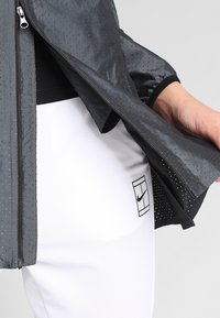 Chaqueta negra perforada con cierre de cremallera y puños elásticos, combinada con pantalones blancos que presentan un logo de Nike en negro. Tejido suave y ligero.