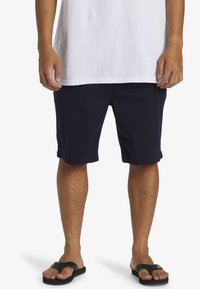 Pantalones cortos de algodón azul marino con un diseño recto, dobladillo con vuelta y bolsillos laterales, combinados con chanclas negras en pies descalzos.