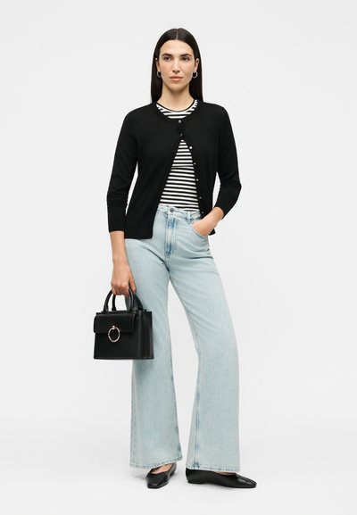 Γυναίκα με μαύρη ζακέτα, μαύρο-άσπρο ριγέ τοπ, ανοιχτό μπλε παντελόνι με φαρδιά πόδια, μαύρα μπαλαρίνια, κρατώντας μαύρη τσάντα με λεπτομέρεια δαχτυλιδιού.