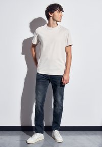 Hombre de pie contra una pared blanca, vistiendo una camiseta básica de color beige claro, jeans oscuros y zapatillas blancas, mirando hacia su derecha.