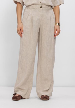 Woman wearing beige wide-leg pants, brown flat shoes, and a beige buttoned shirt, standing against a plain light background.