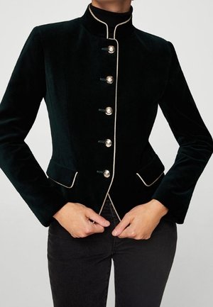 Mujer con chaqueta entallada de terciopelo negro con botones plateados y ribetes claros a lo largo del cuello y los bolsillos, manos cerca de la cremallera.